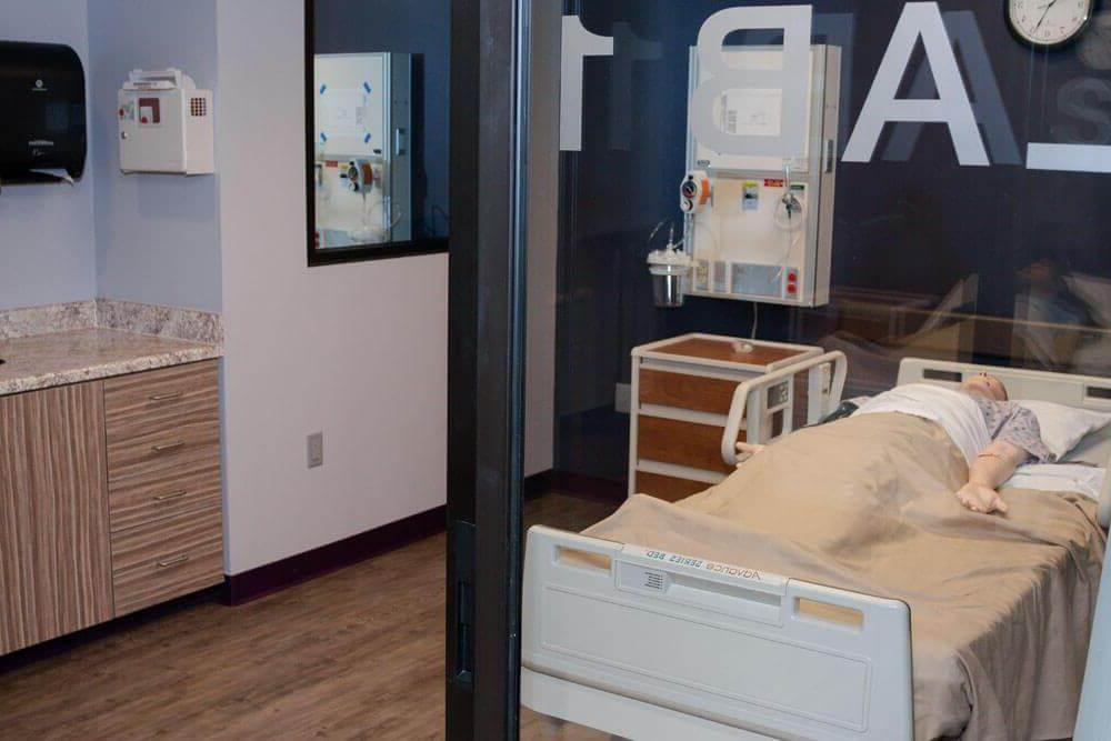 View looking through window of simulation patient room with mannequin laying in hospital bed