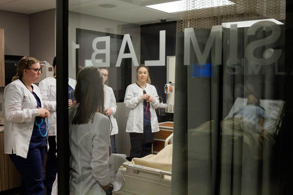 view looking through window into sim lab with professor teaching students